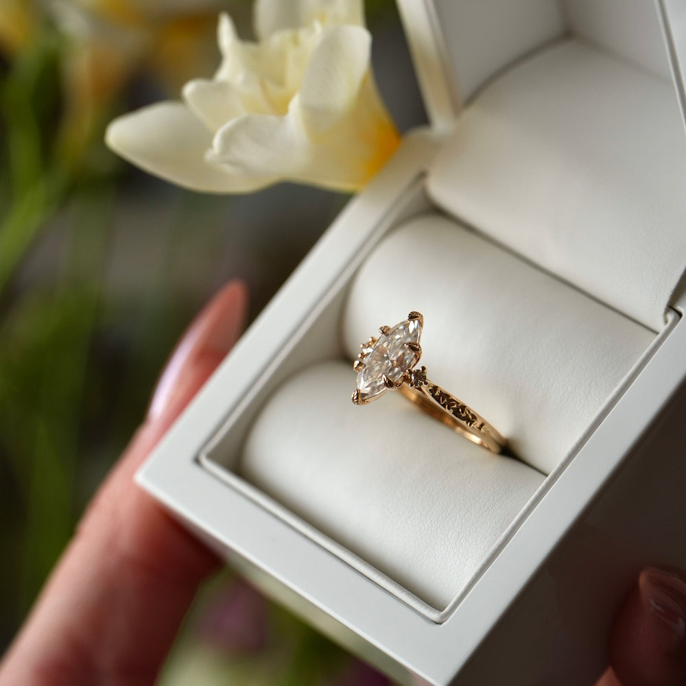 Gold ring with a diamond in a white box held by a hand, with a blurred floral background.