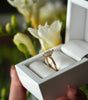 Gold ring with a diamond in a white box held by a hand, with flowers in the background