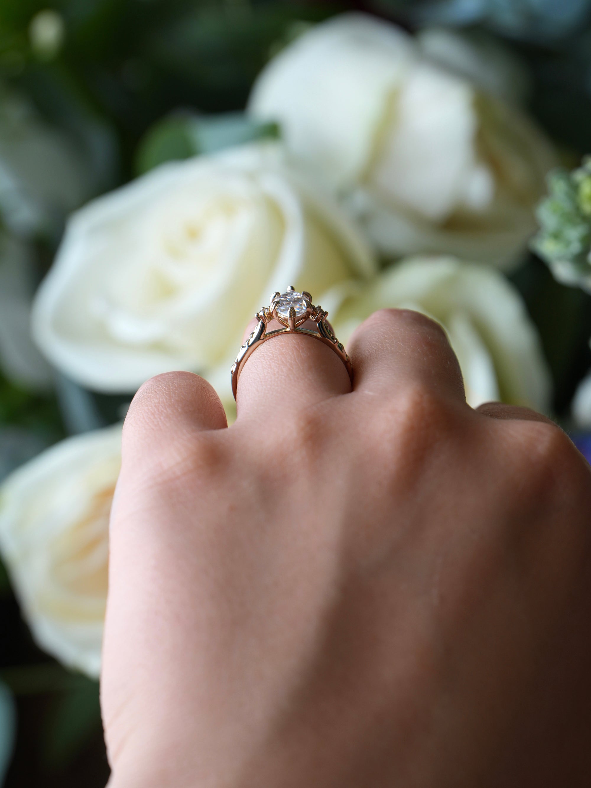 Hand wearing a ring with a diamond, held against a background of white flowers.