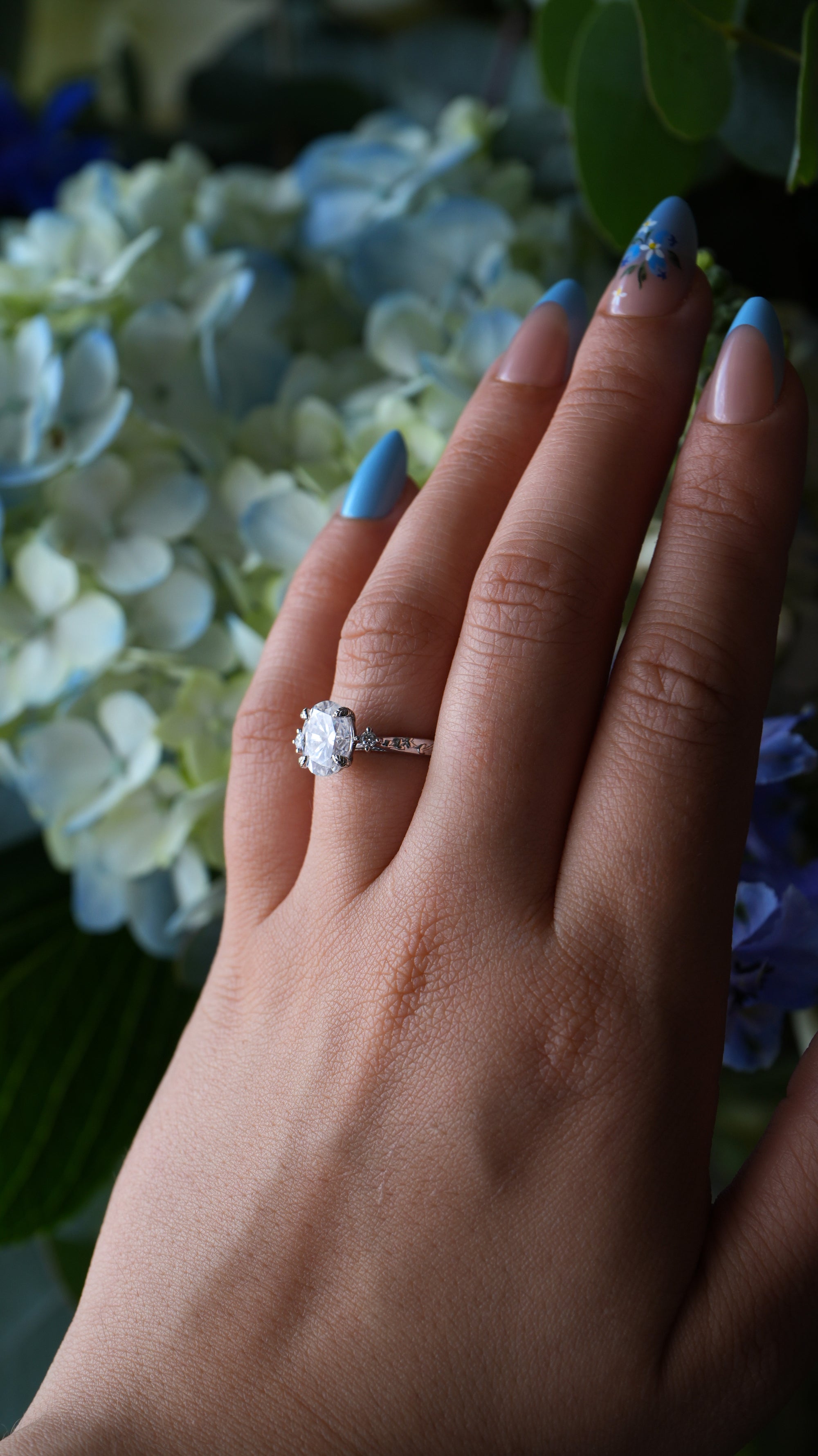 Hand wearing a diamond ring with blue nail polish against a floral background
