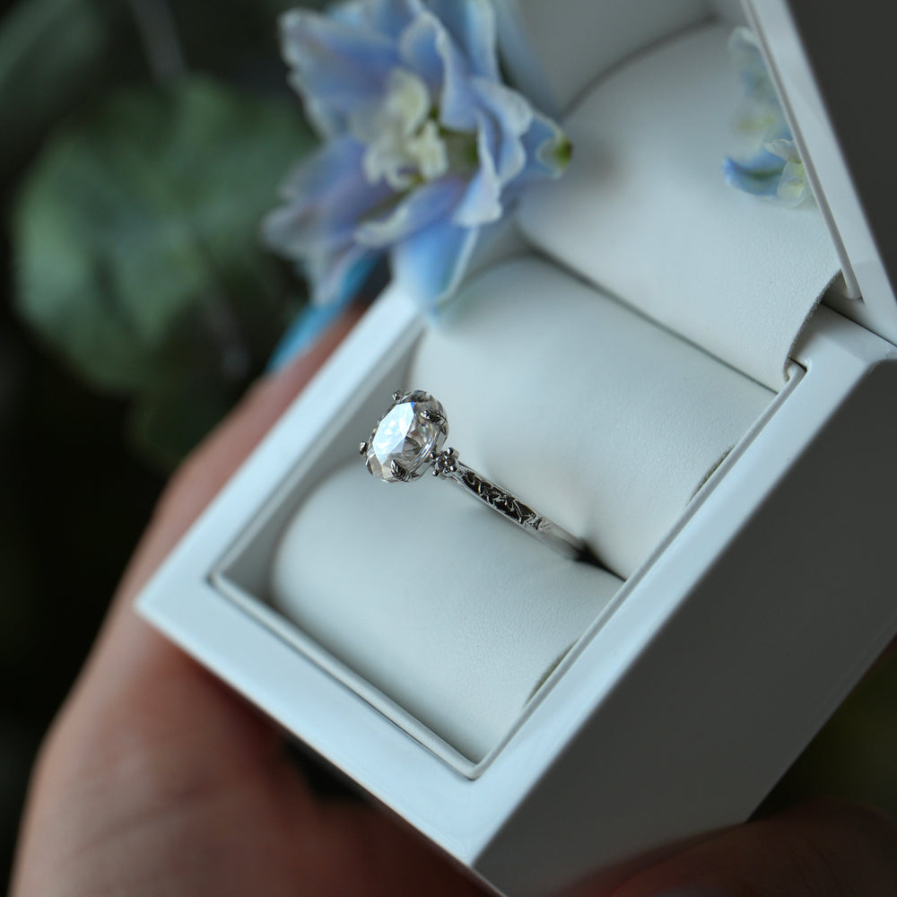 Diamond ring in a white jewelry box held by a hand with floral nail polish.