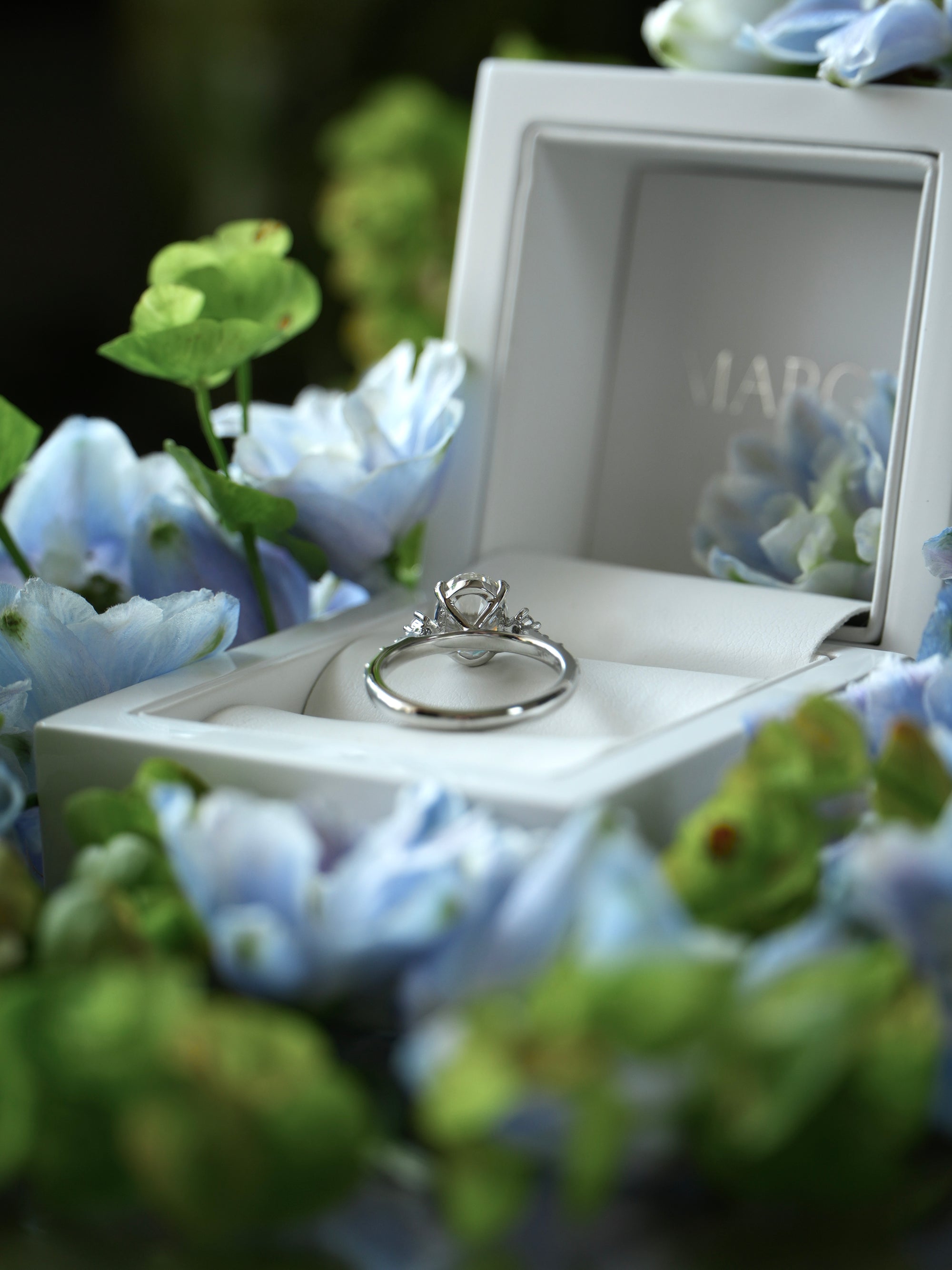 Silver ring in a white box with 'Marc' branding, surrounded by blue flowers.