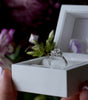 Diamond ring in a white box held by hands with a blurred floral background