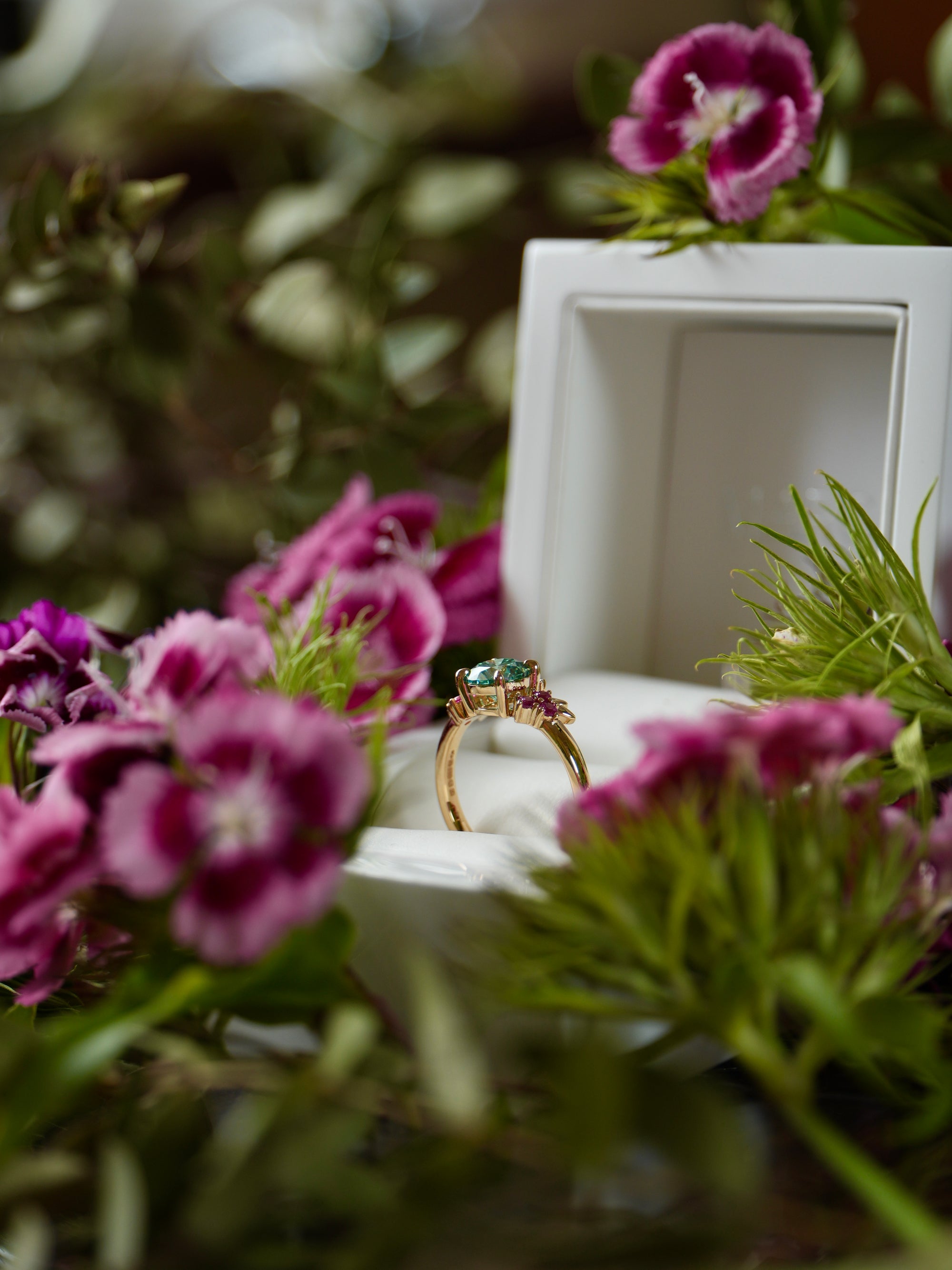 Gold ring with a green gemstone in a white box surrounded by pink flowers.
